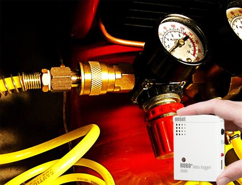 A hand holding a HOBO state data logger beside a compressed air connection with a pressure gauge.